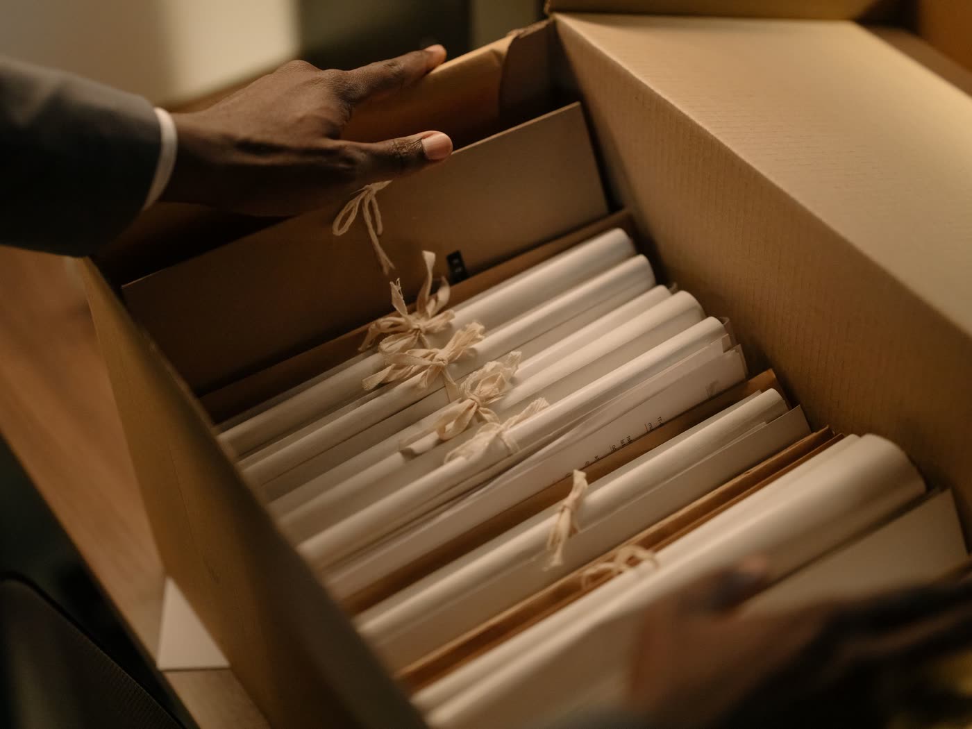 Hands lift a ribbon-tied bundle of paper files out of a cardboard archive box in warm lamplight
